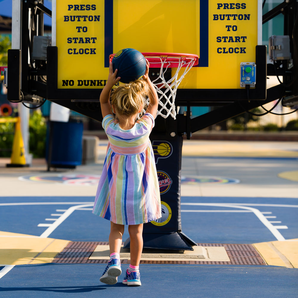 Photo of the back of a girl holding a multicolored basketball over her head and standing in front of a basketball goal.